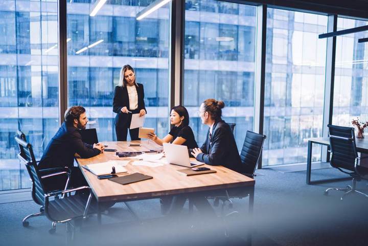 Four business professionals meeting around a conference table in a modern office with large floor-to-ceiling windows, reviewing documents and a tablet while one woman stands presenting.