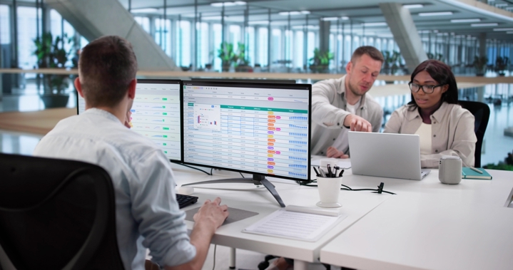 Three banking professionals collaborating at a desk with dual monitors displaying financial data and dashboards, illustrating unified systems replacing disconnected tools.