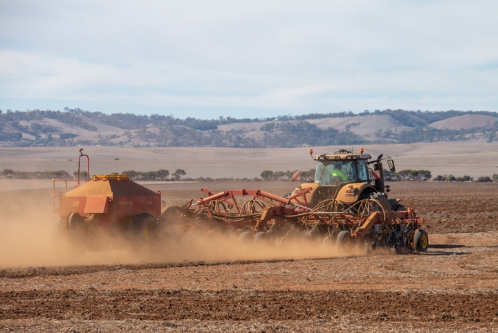 A tractor pulling a seeding machine across a dry Australian paddock, kicking up dust against a backdrop of rolling hills.