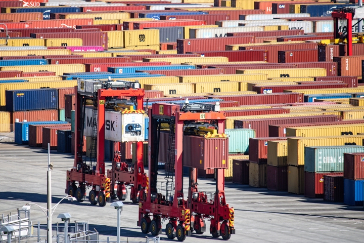 Large straddle carriers moving shipping containers across a busy international port terminal, with rows of colourful containers from global freight operators stretching into the background.