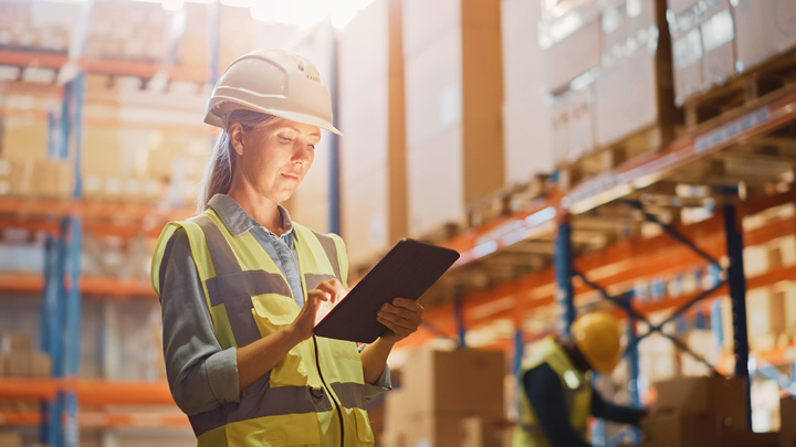 A person in a safety vest and hardhat using a tablet in a wharehouse