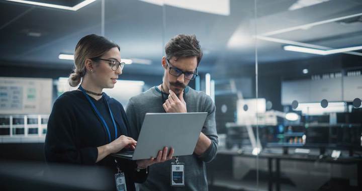 Two professionals wearing ID badges review information on a laptop in a modern office, standing in front of glass walls with a focused, thoughtful expression.