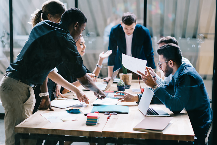 Several people gather around a table in an office. They are looking at laptop and papers in front of them and in the middle of the table, brainstorming for a new product.