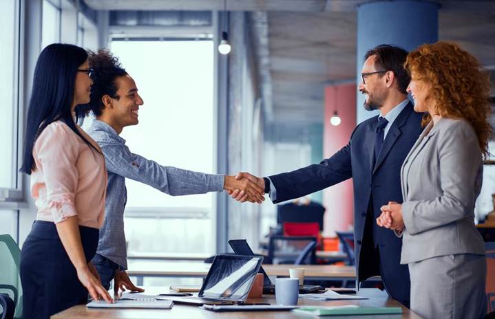 Two business professionals shaking hands across a desk in a modern office while two colleagues stand beside them, smiling, with a laptop and documents on the table.