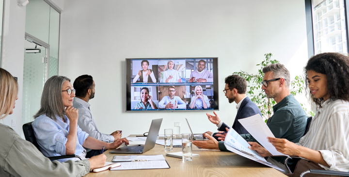 A group of business professionals sit around a conference table reviewing documents while participating in a video meeting displayed on a screen in a modern office.