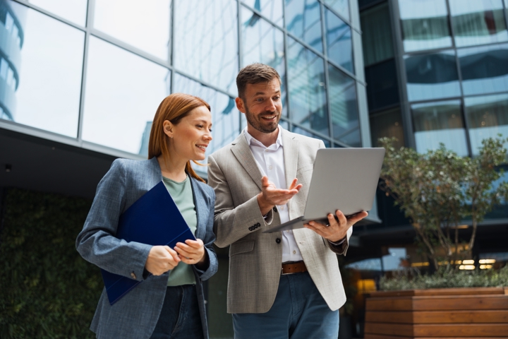 Two business professionals standing outdoors in front of a modern glass office building, smiling and looking at a laptop screen together. The woman holds a blue folder; the man gestures toward the laptop he is holding.