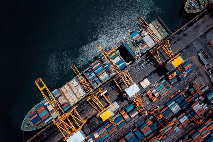 “Top-down view of a container port with cranes and stacked shipping containers alongside a cargo vessel.”