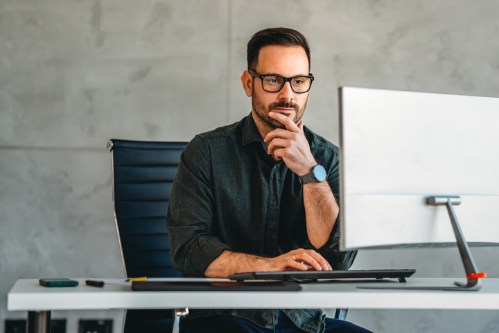 A banker sitting at a desk, looking at a computer monitor while thinking, with one hand resting on his chin.