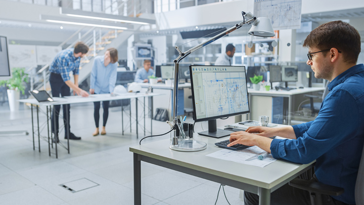 An engineer works on the design for a prototype on his computer. His colleagues are discussing the project in the background and looking over plans..