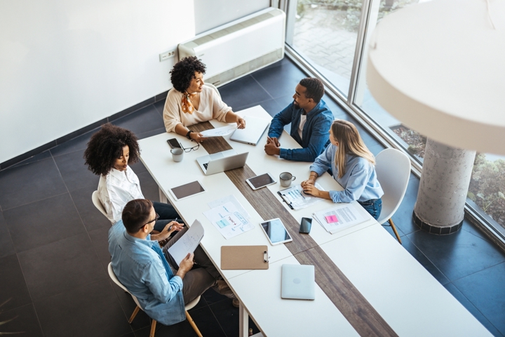 A group of colleagues meet around a table with laptops and printed documents, discussing industry information and aligning assumptions across teams.