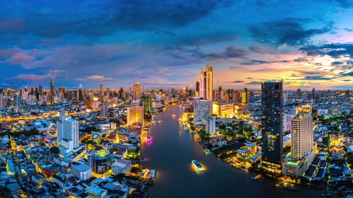“Nighttime city skyline along a river in Bangkok, showing dense urban development and illuminated commercial buildings.”