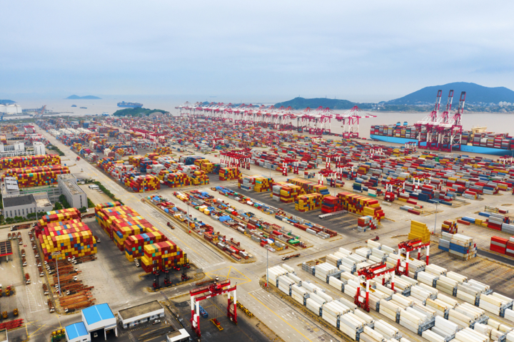 “Aerial view of a major container terminal with stacked shipping containers, cargo cranes and freight vehicles, illustrating global trade flows.”