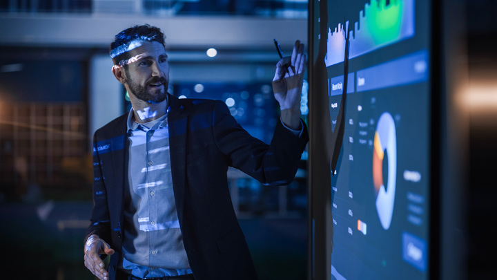 A business leader presents detailed data to his team. He is standing in front of a screen that shows data visualizations and pointing to a chart.