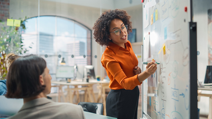 Two people watch a trusted advisor write a plan on a whiteboard. They are in a bright, modern office with large windows.