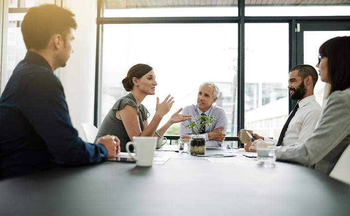 alt text for this image 10:41 AM A group of five business professionals engaged in a discussion around a conference table in a bright, modern office meeting room.