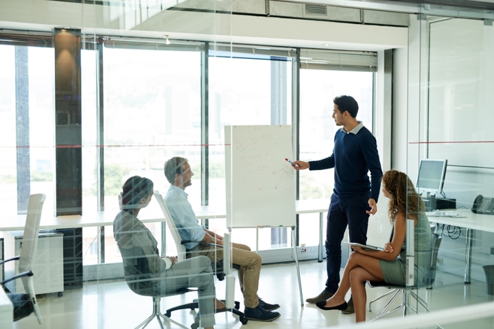Business professional presenting to three colleagues in a glass meeting room, pointing to a whiteboard while the group listens, representing discussion without immediate action.
