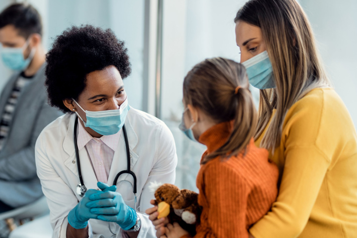 A pediatrician wearing a white coat and stethoscope talks to a child and parent.