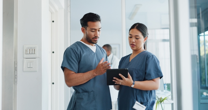 Two healthcare workers wearing blue scrubs look at a tablet, discussing a patient's results.