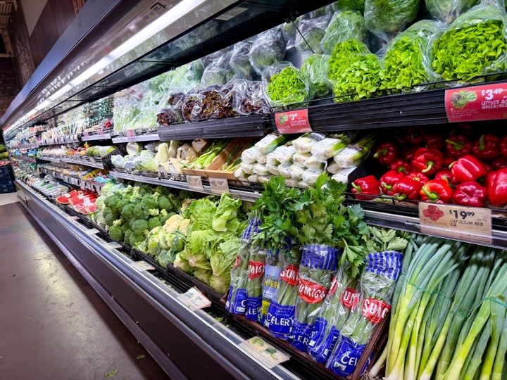 A fully stocked fresh produce aisle in an Australian supermarket, displaying vegetables including broccoli, celery, capsicum and lettuce with price tags visible.