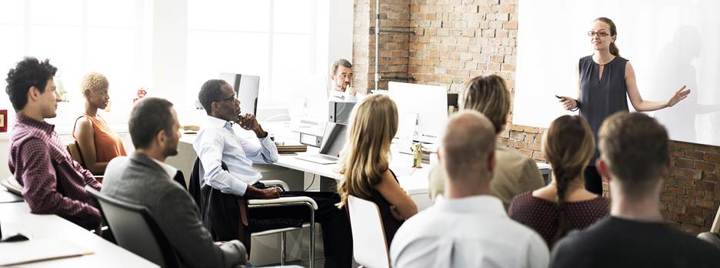 A woman giving a presentation to a seated group of colleagues in a bright office with exposed brick walls, as attendees listen and face a projected screen.