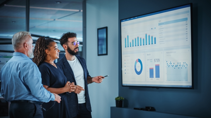 Three people participate in a strategy meeting. They are standing in front of a screen that shows a range of charts and data. One is holding a remote and discussing the data, while the other two listen.