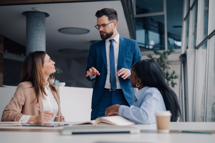 Three business professionals discuss data around a table in a modern office, with one colleague standing and explaining while others review notes.