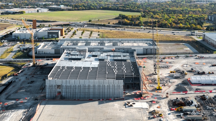 Aerial view of a large data centre under construction on the edge of a city, with two tower cranes, exposed steel roof framing, concrete tilt panels and cleared earth across the site, bordered by a motorway and open grassland.