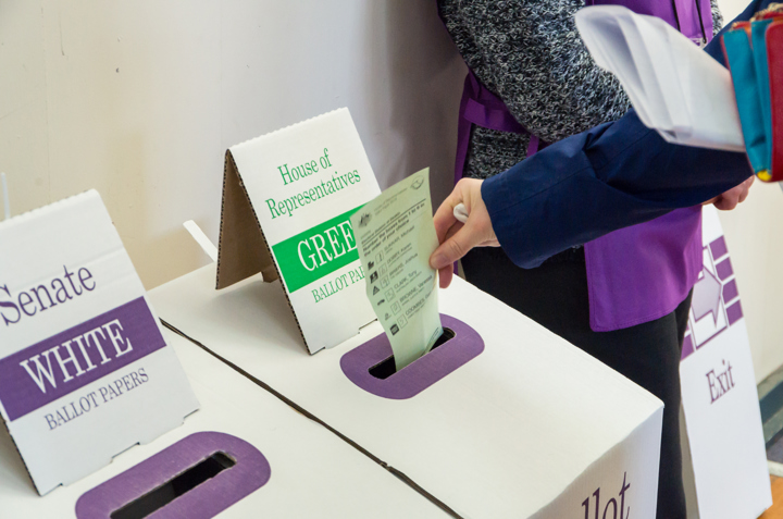 A voter placing a green House of Representatives ballot paper into a cardboard ballot box at an Australian polling station, with signage for Senate and House of Representatives papers.