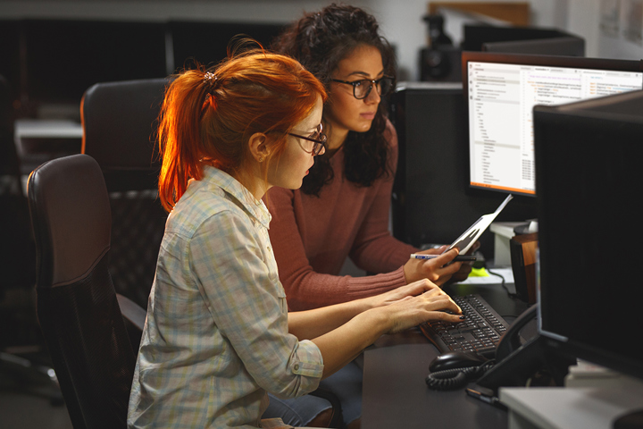 A software developer sits in front of a computer at a row of desks. The developer is typing, and code is visible on one of the monitors in front of them. Another person sits beside them, looking at the screen.