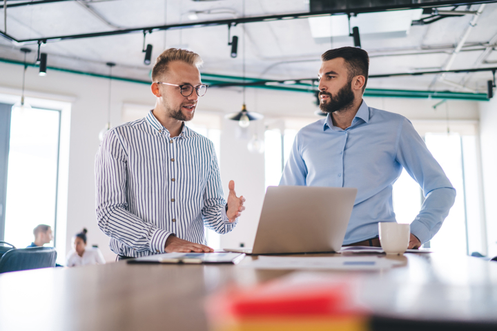 Two professionals standing at a high table in a modern office, discussing work while looking at a laptop.